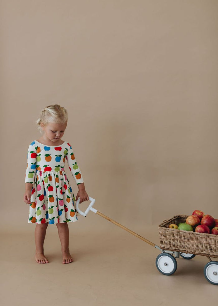 Child in a colorful dress pulling a cart with fruits against a beige background