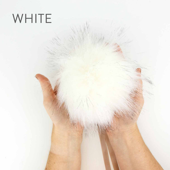 Hands holding a 6-inch faux fur pom pom in White with black-tipped fibers and a ribbon tie showing the full fluffy size