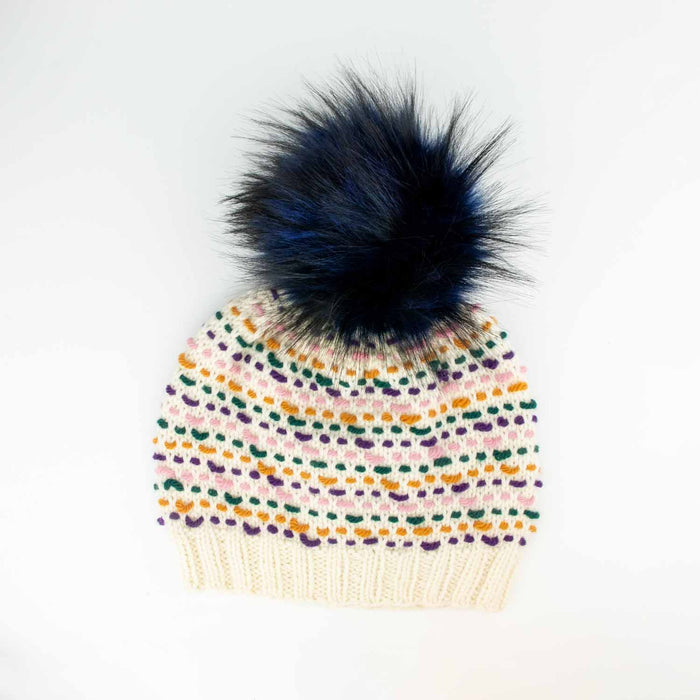 Colorwork knitted hat in cream with pink, green, purple, and ochre stitches topped with a Midnight Blue 6-inch faux fur pom pom on a white background