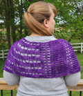 Back view of a model wearing the Galliano Revenge Wrap in purple African Violet yarn draped over the shoulders, showing the open lacework grid pattern with alternating checked and mesh sections, worn over a gray sweater outdoors on a deck with green trees and a white fence