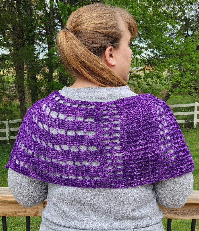 Back view of a model wearing the Galliano Revenge Wrap in purple African Violet yarn draped over the shoulders, showing the open lacework grid pattern with alternating checked and mesh sections, worn over a gray sweater outdoors on a deck with green trees and a white fence