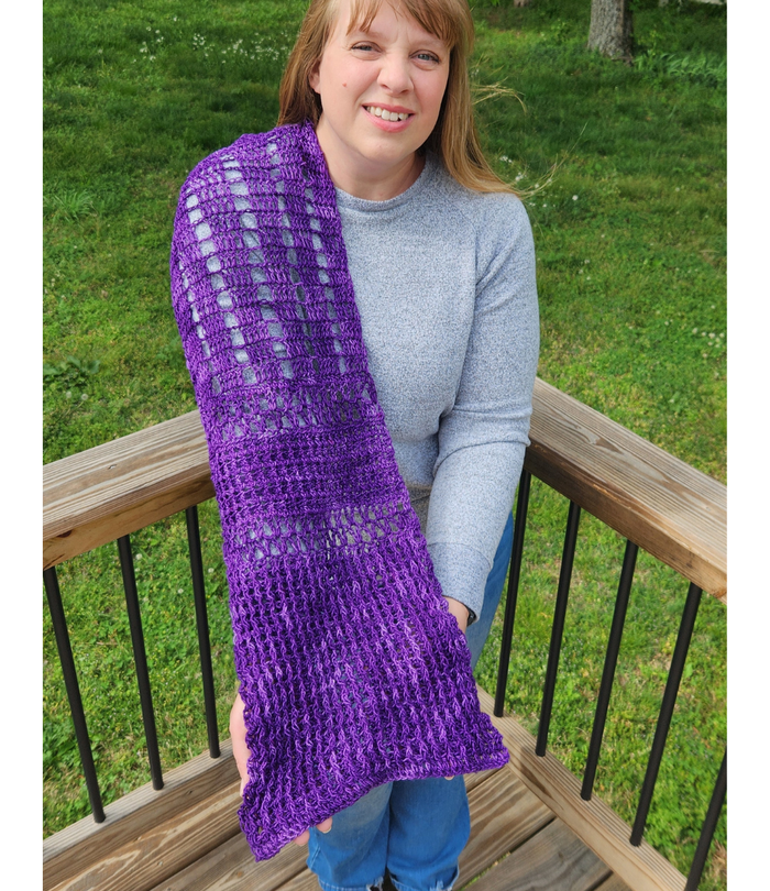 Front view of a smiling model holding the Galliano Revenge Wrap open to display its full length, showing the purple lace crochet wrap draped over one shoulder with geometric grid and V-stitch sections visible, modeled outdoors on a wooden deck