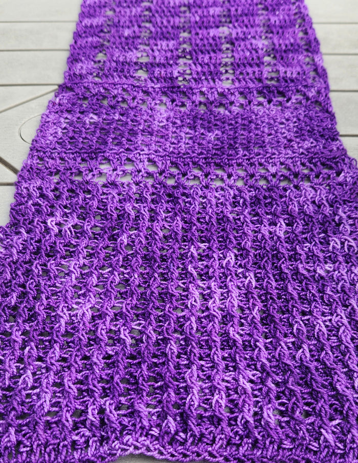 Close-up detail of the Galliano Revenge Wrap laid flat on a gray wood surface, showing the crochet stitch pattern with alternating sections of open lacework grid and denser V-stitch rows in variegated purple African Violet yarn