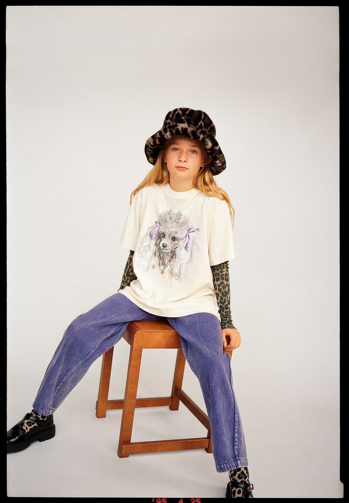 Child sitting on a stool wearing a graphic t-shirt, jeans, and a hat with a white background