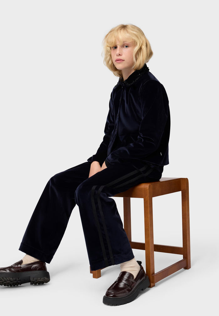 Person wearing a blue velvet outfit sitting on a wooden stool against a white background