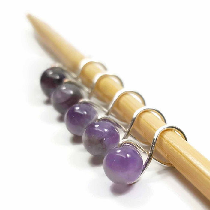 Five amethyst purple gemstone infinity ring stitch markers with silver-toned wire loops displayed on a bamboo knitting needle against a white background