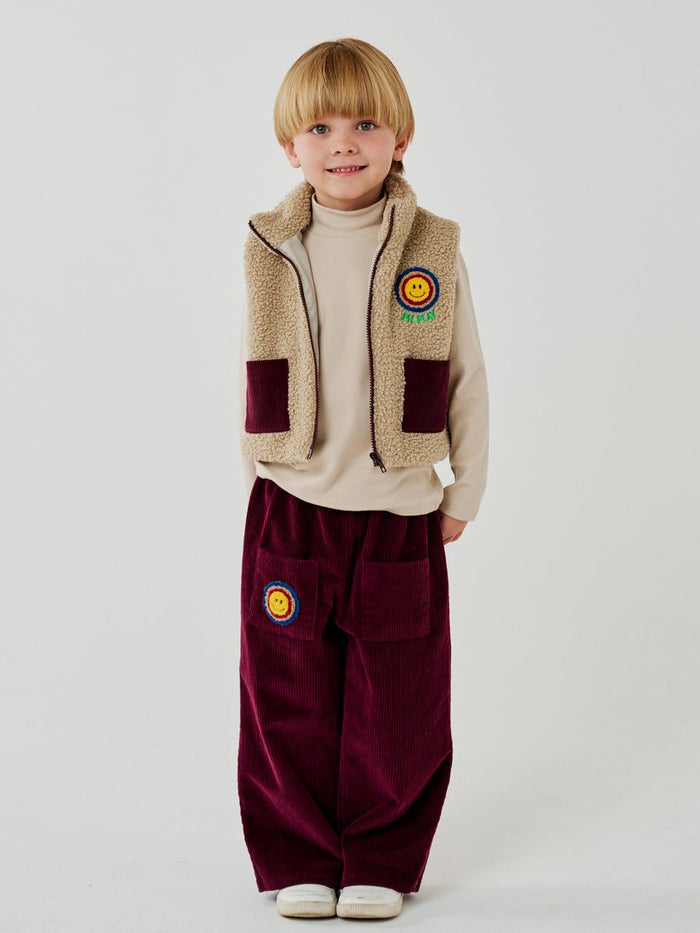 Child wearing a fleece vest and burgundy pants with patches on a white background
