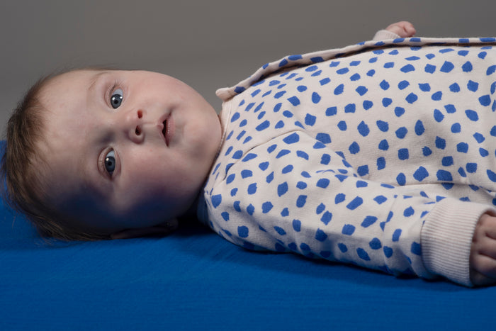A baby lying on a blue surface wearing a white long-sleeve romper with blue spots, photographed in a studio setting.