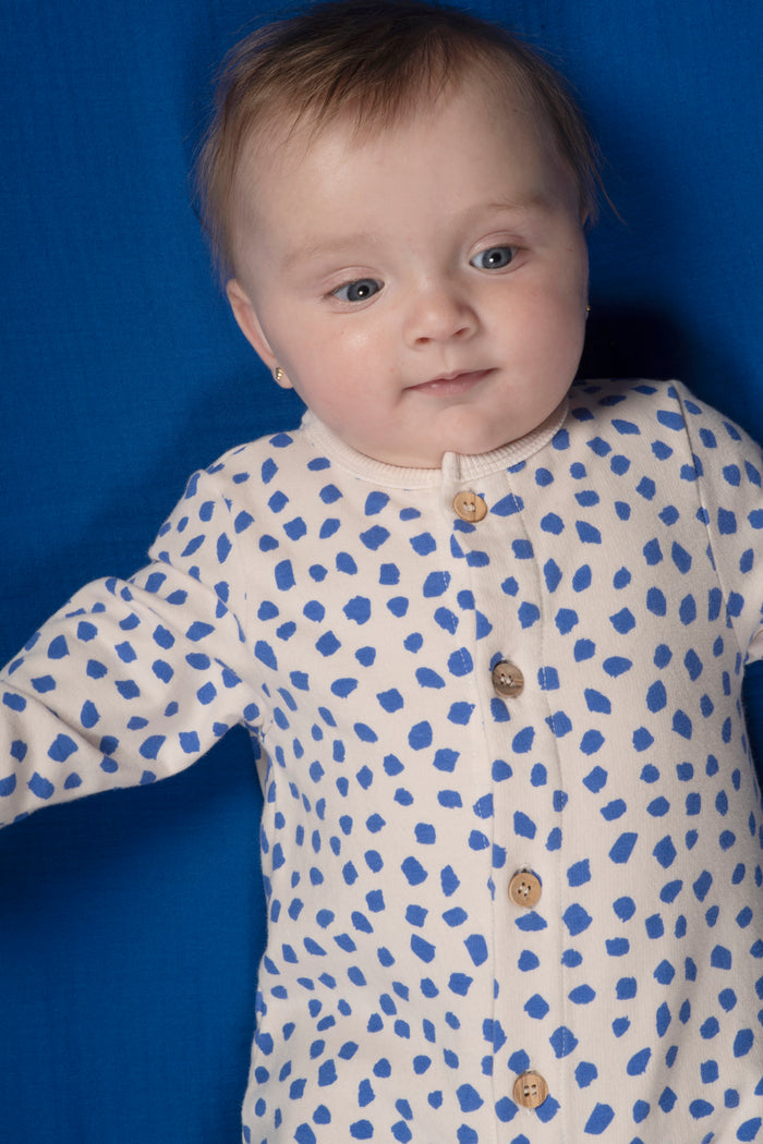 A baby wearing a long-sleeve white footless onesie with blue polka dots and brown buttons, photographed against a solid blue background.