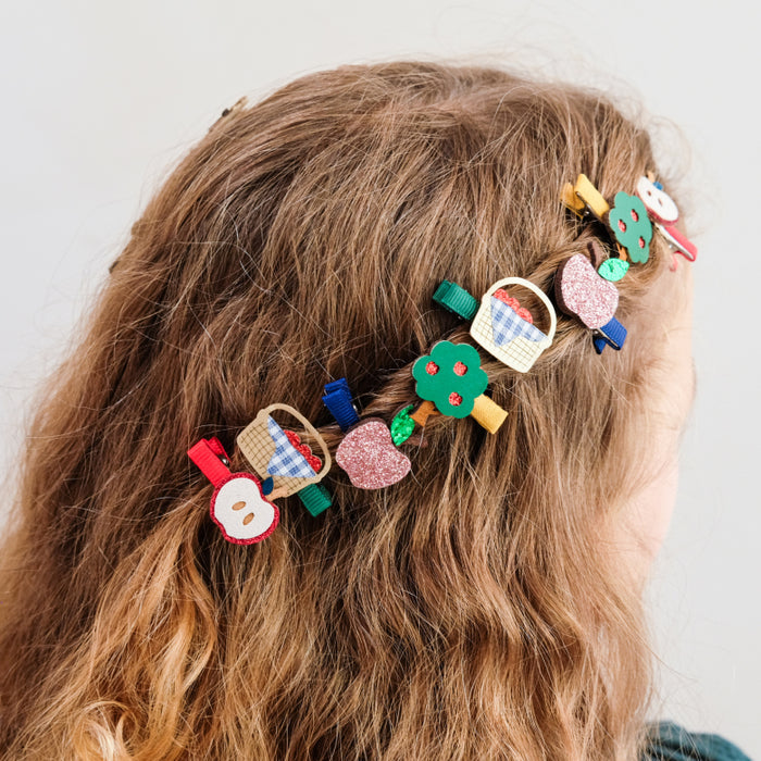 A close-up of a person's wavy light brown hair adorned with multiple colorful decorative hair clips, including basket, apple, and tree designs, against a plain light gray background.