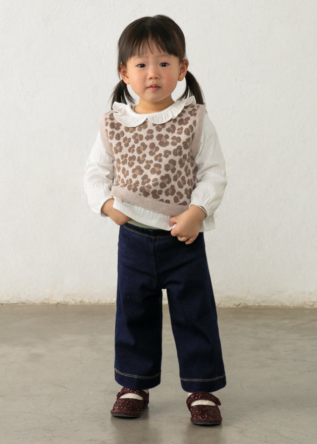 Young girl wearing a leopard print vest over a white shirt with dark blue pants against a plain background