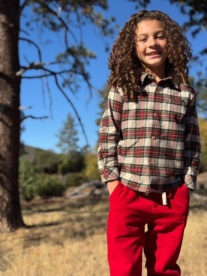 Child wearing a plaid shirt and red pants standing outdoors with trees and blue sky in the background