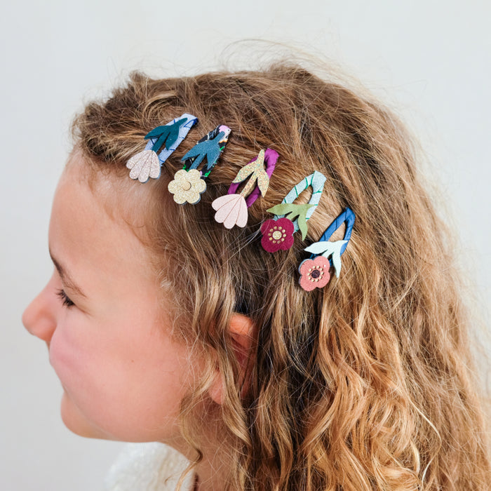 A side-profile view of a young girl with wavy light brown hair wearing a set of colorful fabric flower hair clips against a plain light gray background.