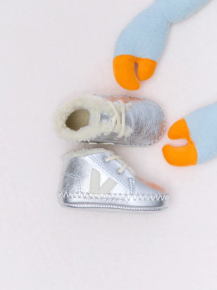 A pair of metallic silver baby booties with white faux-fur trim and white laces, arranged on a light pink background with a blue and orange plush toy partially visible in the corner.