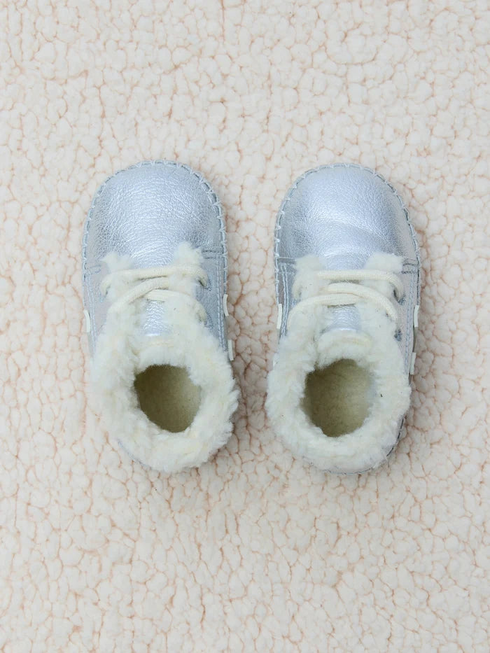A pair of silver metallic baby booties with white faux-fur lining and white laces, laid flat on a textured cream fleece background.