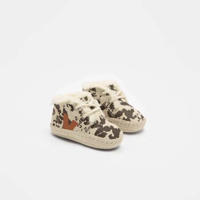 A pair of baby lace-up booties featuring a brown and beige cow print pattern, cream faux fur lining, and a small brown leather logo patch on the side, set against a plain white background.