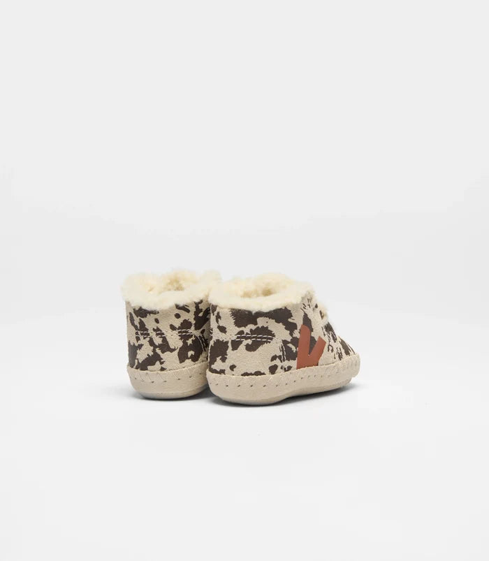 A pair of baby slip-on shoes with a beige and brown cow print pattern, featuring a soft cream sherpa lining and a small brown logo on the side, photographed against a plain white background.