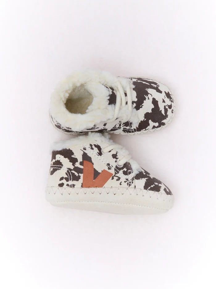 A pair of baby booties featuring a brown and white cow print pattern, white faux fur trim, and a brown leather V-shaped accent on the side, displayed on a clean white background.