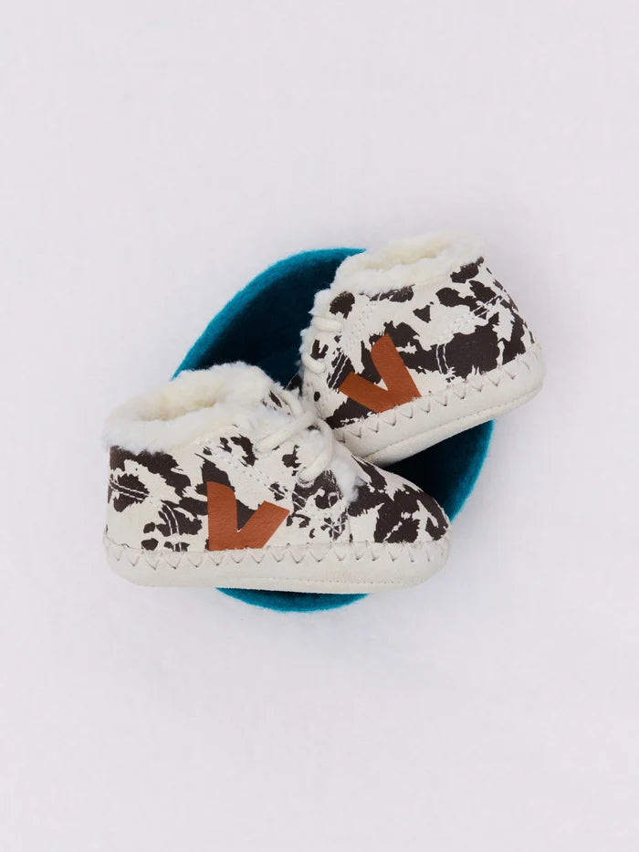 A pair of baby booties with a brown and white cow print pattern, featuring a white faux-fur collar and a brown leather V-shaped logo on the side, resting on a teal circular stand against a white background.