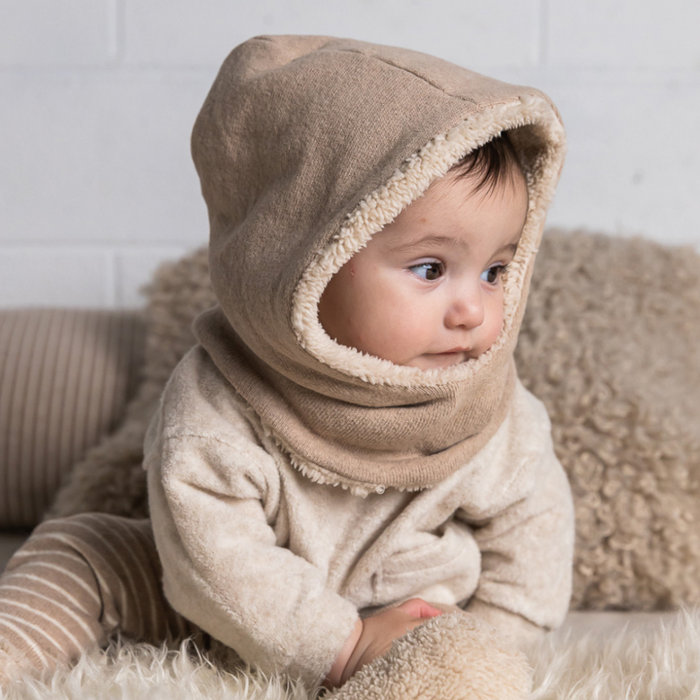 Baby wearing a beige hooded outfit sitting on a soft surface.