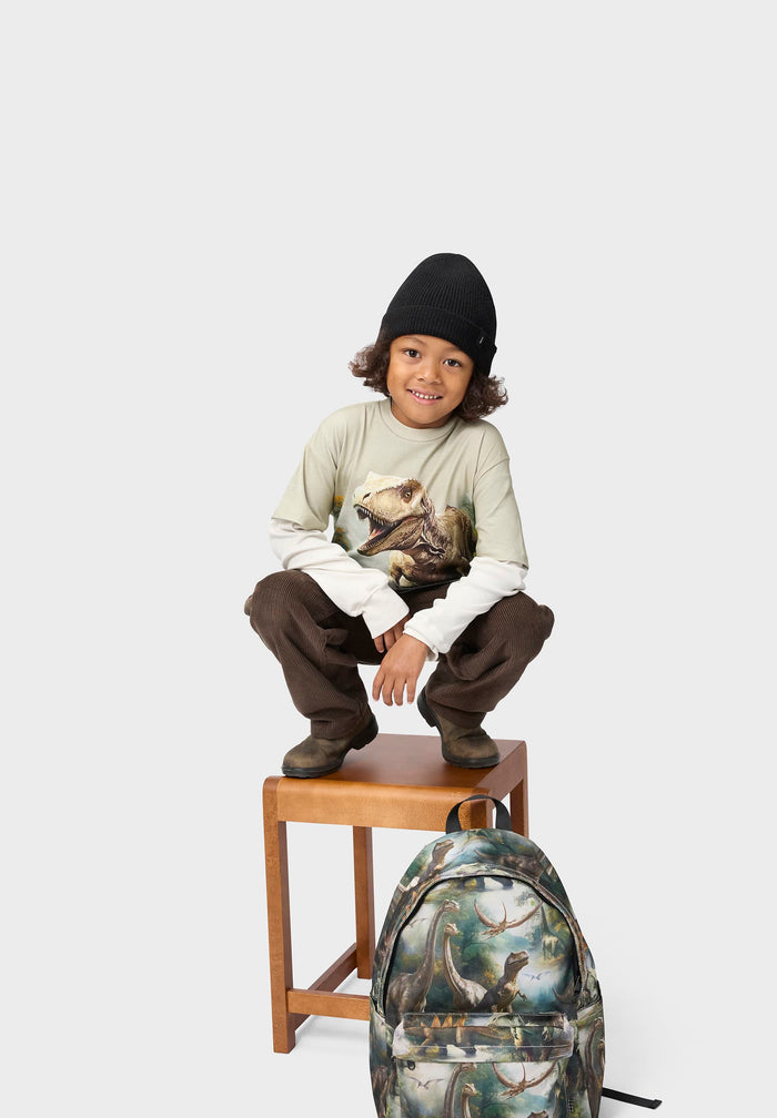 Child sitting on a stool wearing a graphic t-shirt and brown pants, with a backpack featuring a similar design.