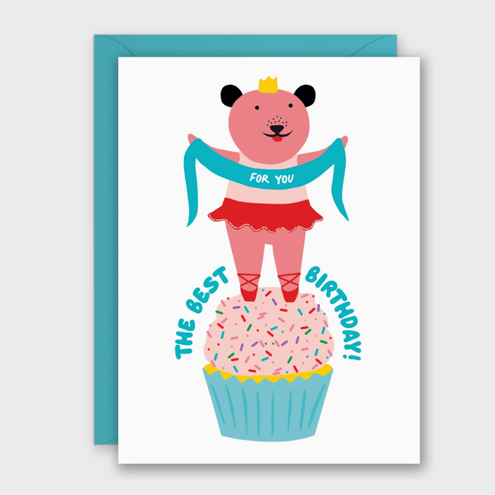 A white greeting card with a turquoise envelope behind it, featuring a cartoon pink bear dressed as a ballerina standing on a cupcake and holding a turquoise banner that reads 'FOR YOU'. The card also says 'THE BEST BIRTHDAY!' in turquoise text.