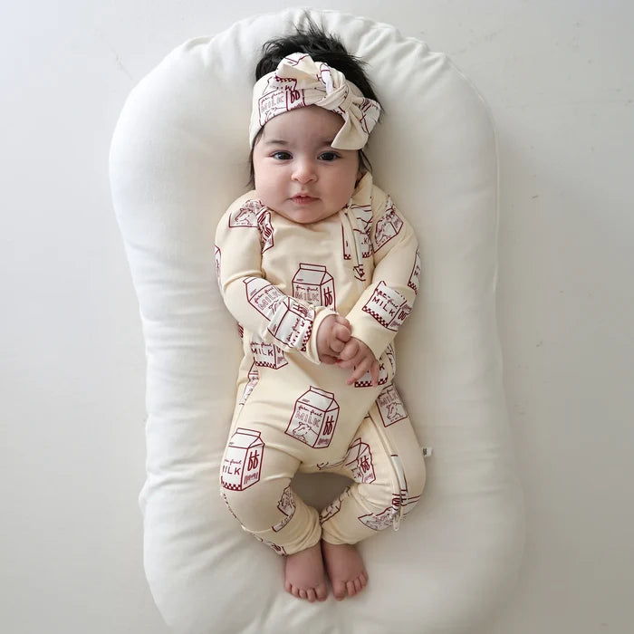 A baby lying on a white curved baby lounger, wearing a cream-colored long-sleeve footless onesie with a red milk carton pattern and a matching headband.