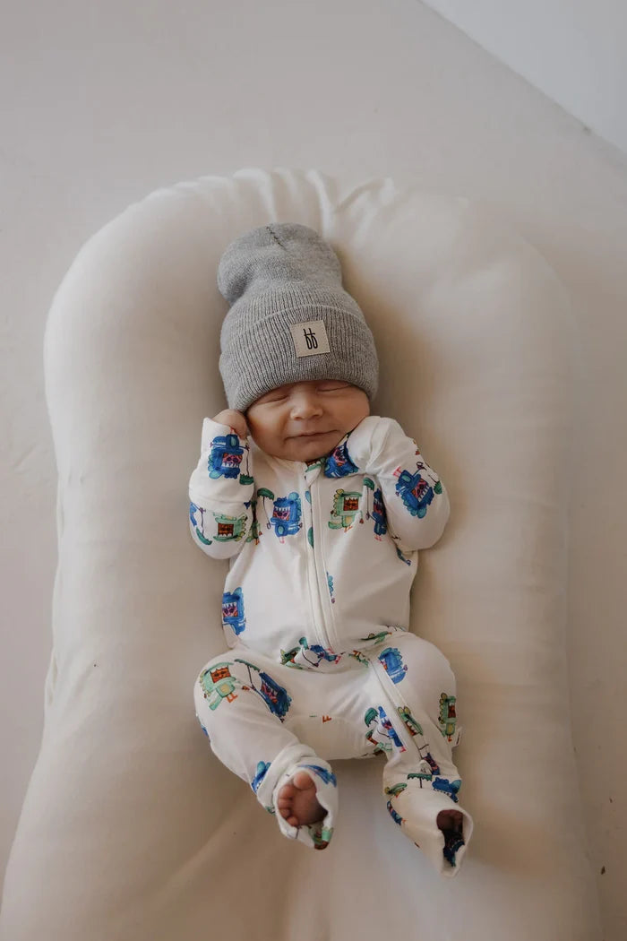 A sleeping newborn baby wearing a white footed onesie with colorful monster prints and a gray knit beanie hat, lying on a white baby lounger.