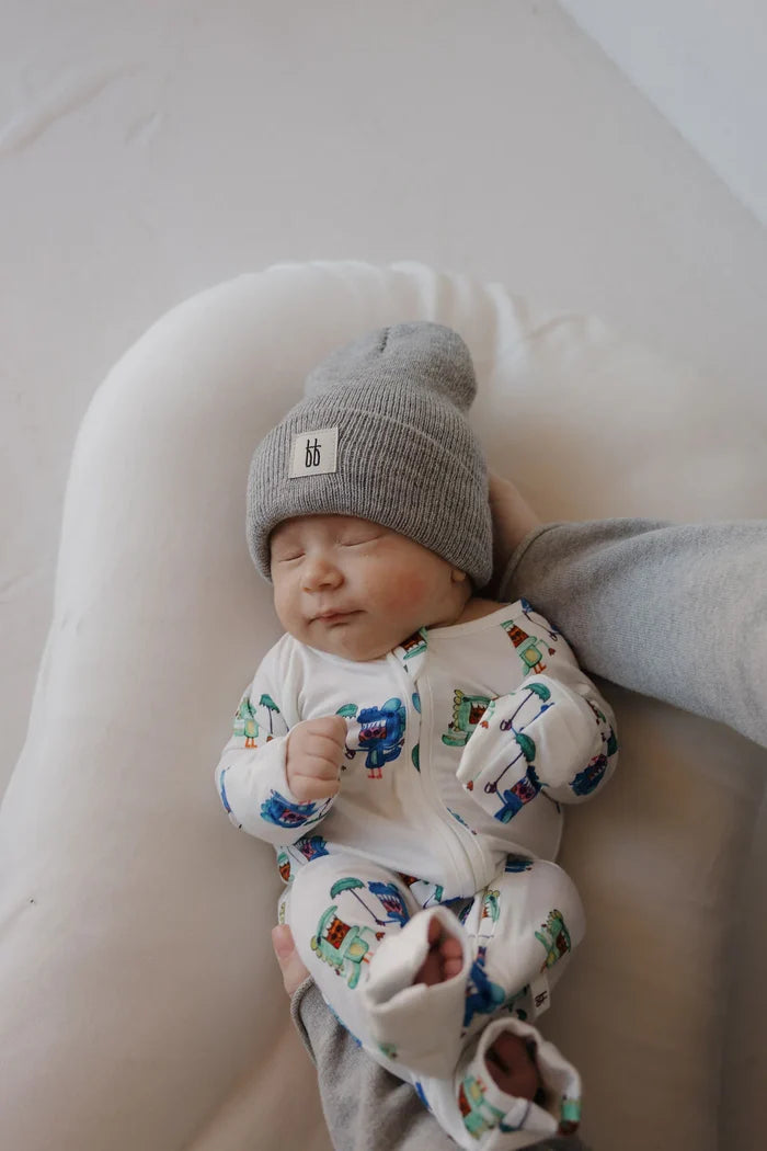 Baby wearing a patterned onesie and gray beanie lying on a white surface.