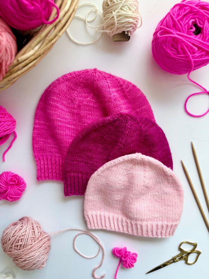 Three Base Doodle Beanie 1 hats in pink shades with yarn and needles