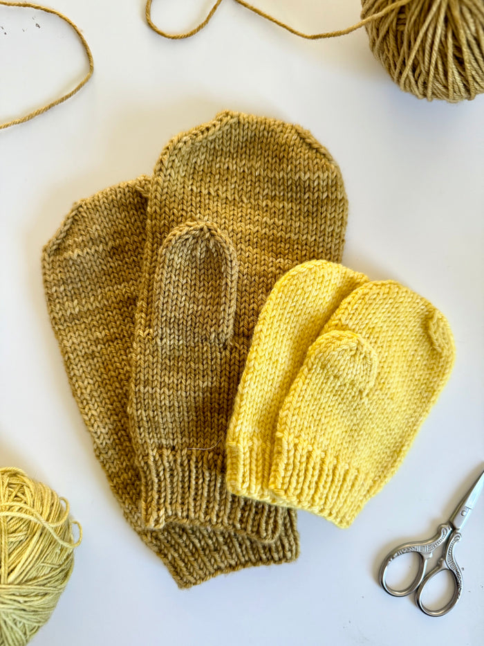 Two pairs of Base Doodle Mittens in olive and yellow with yarn and scissors