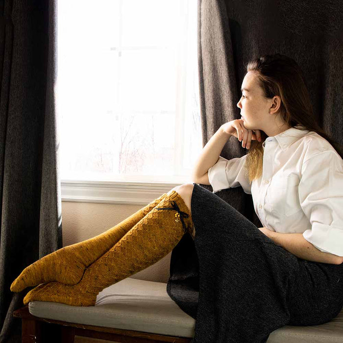 Model sitting on a window bench wearing hand-knit Bees Knees knee-high socks in golden honey tweed yarn with honeycomb lace pattern and black ribbon bow with gold honeycomb and bee charms