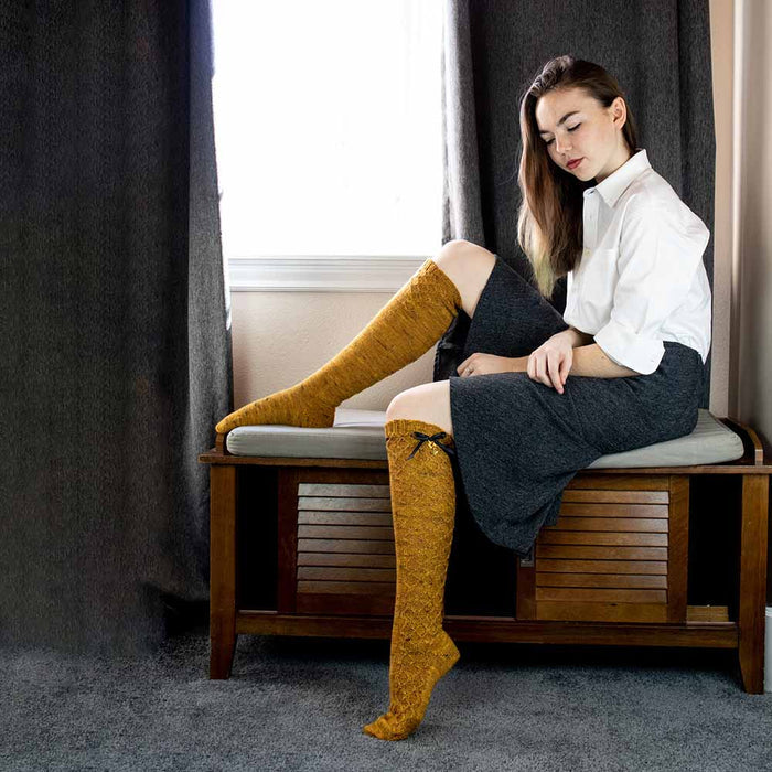 Model sitting on a window bench looking down at hand-knit Bees Knees knee-high socks in golden honey tweed yarn with honeycomb lace texture and black satin ribbon bows