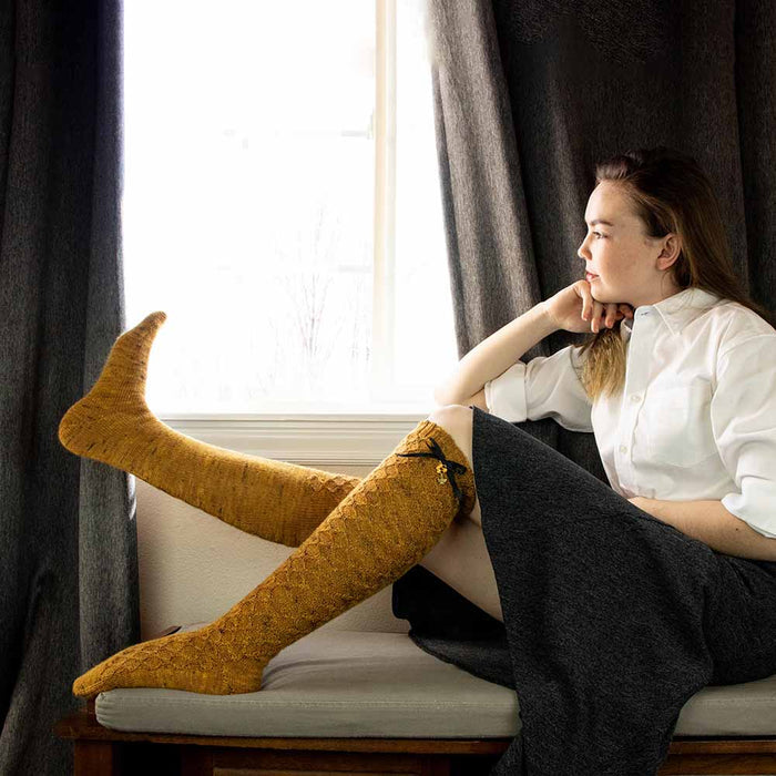 Model sitting on a window bench gazing out the window while wearing Bees Knees knee-high socks in golden honey tweed yarn with legs extended showing the honeycomb lace pattern and black ribbon bow accents