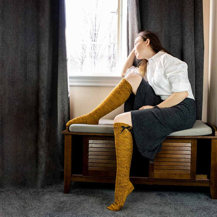 Model leaning against a window seat with one leg raised showing the full length of the Bees Knees knee-high sock in golden honey tweed yarn with honeycomb lace and black ribbon bow