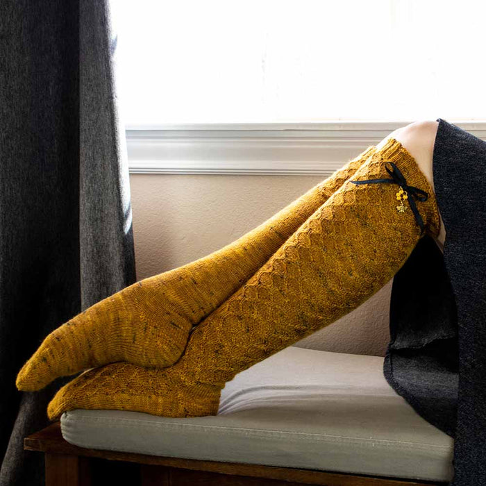 Close-up of both feet resting on a cushion showing the Bees Knees knee-high socks in golden honey tweed yarn with honeycomb lace pattern and black ribbon bows with gold honeycomb and bee charms