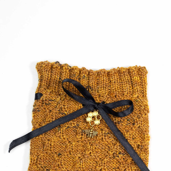 Close-up of the Bees Knees sock cuff showing the ribbed top edge, golden honey tweed honeycomb lace stitch pattern, and black satin ribbon bow with gold honeycomb and bee charms