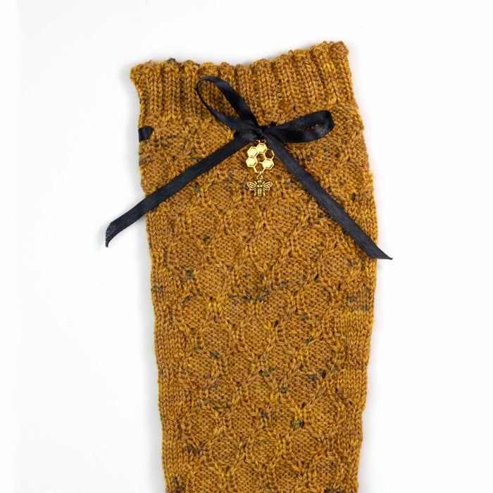 Flat lay close-up of the Bees Knees sock showing the honeycomb lace pattern in golden honey tweed yarn with a black satin ribbon bow and gold honeycomb and bee charm detail at the cuff