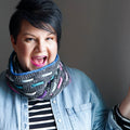 Model wearing the Bennet Cowl in dark gray with colorful woven cord accents in blue, purple, teal, and pink over a denim jacket and striped shirt