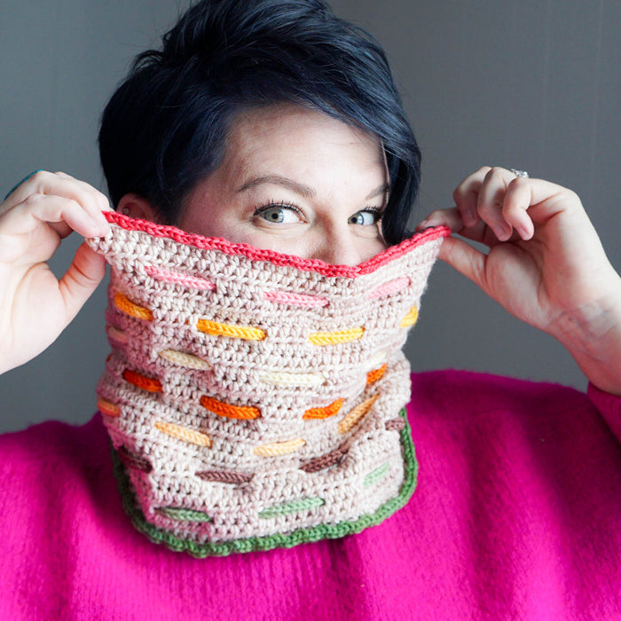 Model playfully pulling up a Bennet Cowl in beige with warm-toned woven cord accents in coral, pink, orange, yellow, brown, and green worn over a bright pink sweater