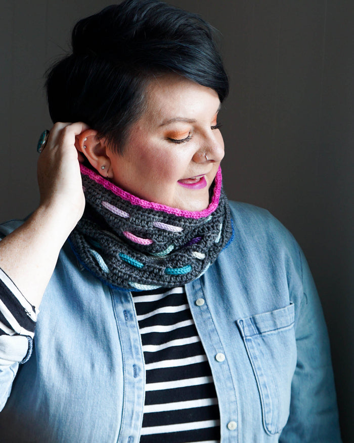Model wearing the Bennet Cowl in dark gray with pink, purple, teal, and pastel woven cord accents and a bright magenta trim at the top styled with a denim jacket