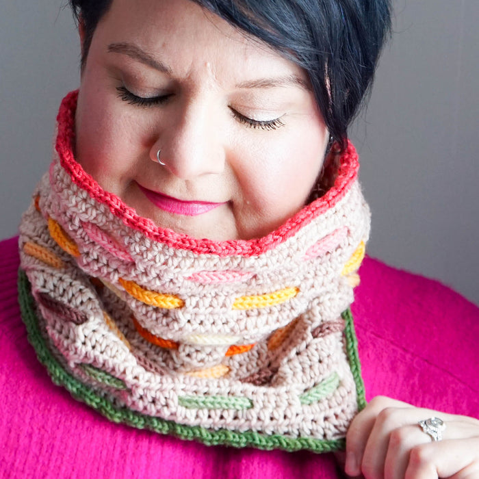 Close-up of model wearing the Bennet Cowl in beige with warm coral, orange, yellow, and green woven cord accents pulled up high over a bright pink sweater