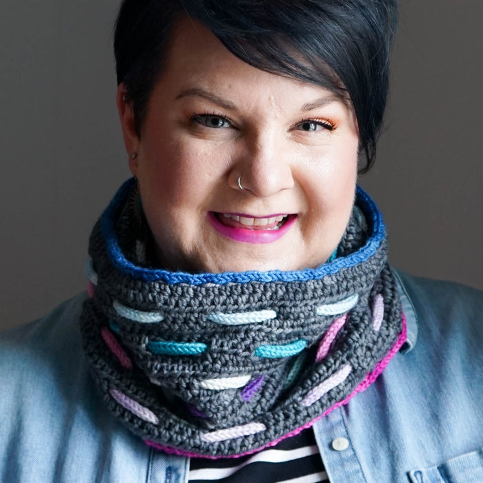Model smiling and wearing the Bennet Cowl in dark gray with blue trim and colorful woven cord accents in blue, teal, pink, lavender, and mint over a denim jacket and striped shirt