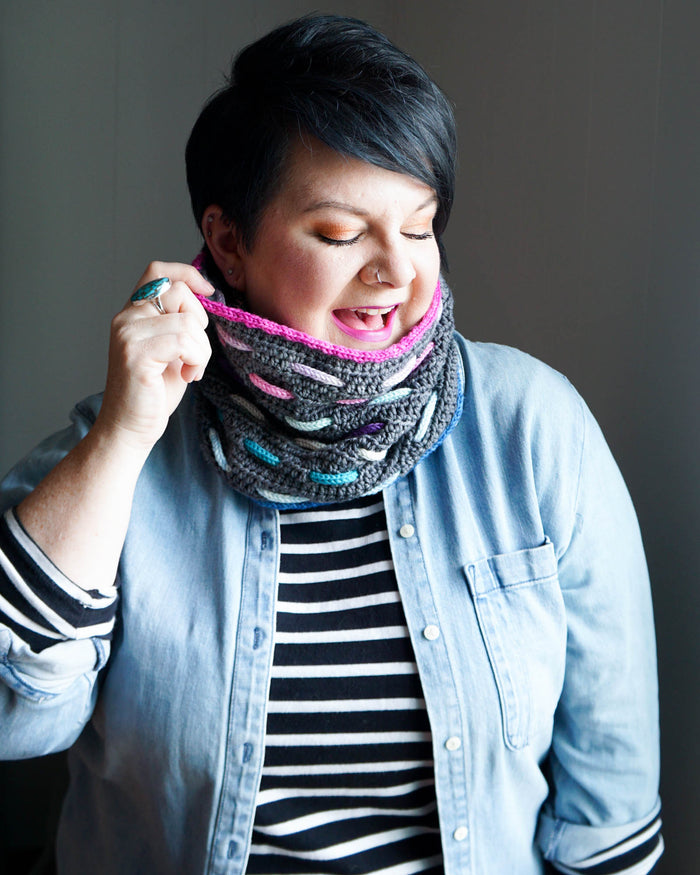 Model wearing the Bennet Cowl in dark gray with pink trim and woven cord accents in teal, pink, lavender, and blue, tilting head to one side with a denim jacket