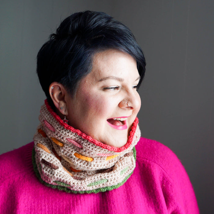 Model laughing with arm raised while wearing the Bennet Cowl in dark gray with blue trim and colorful woven cord accents over a denim jacket and striped shirt