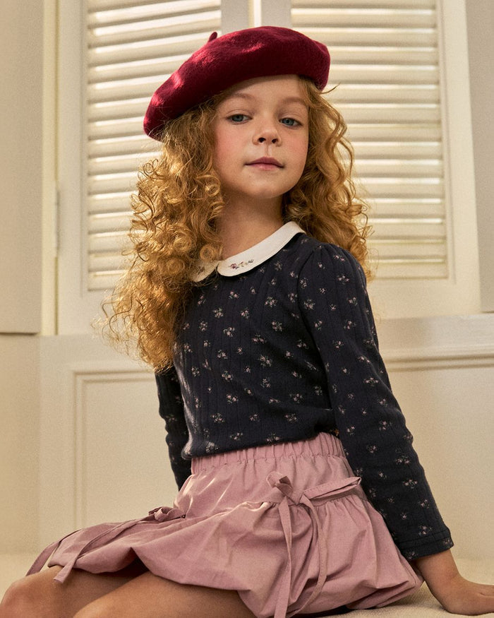 Young girl wearing a wine color beret and dark blue top with pink skirt sitting in front of white shutters.