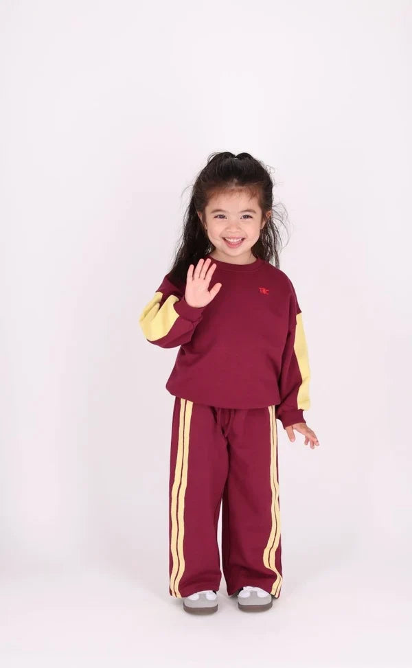 child wearing burgundy wide leg sweatpants with drawstring, 2 pale yellow side stripes and matching burgundy sweatshirt with yellow colorblocking on sleeves with white background
