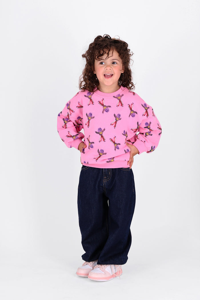 A young child with curly brown hair stands against a plain white studio background, wearing a pink long-sleeve sweatshirt covered in cat print, dark blue jeans, and light pink sneakers.