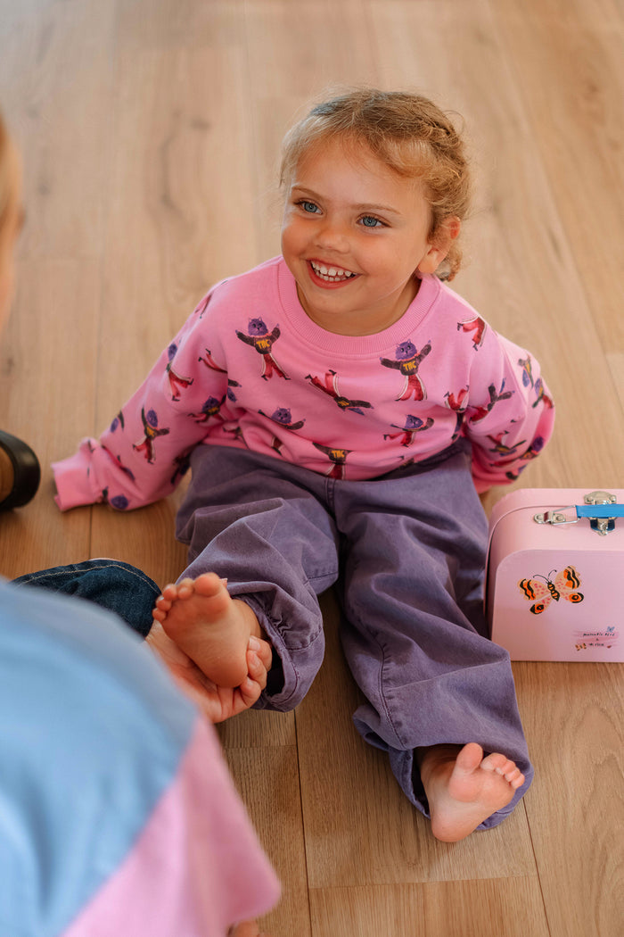 A young child sitting on a light wood floor, wearing a pink long-sleeve shirt with a colorful cat print and purple pants. A small pink lunchbox sits beside them.