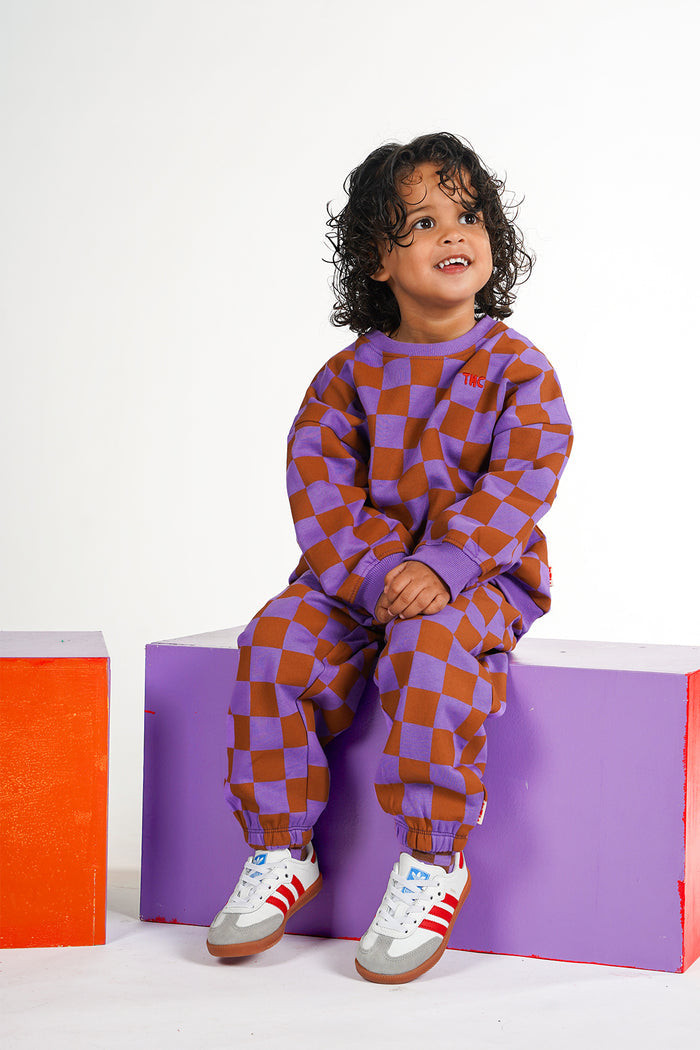 A young child with curly dark hair sits on a purple block wearing a matching purple and brown checkerboard sweatshirt and jogger pants, with white sneakers, against a white studio background.